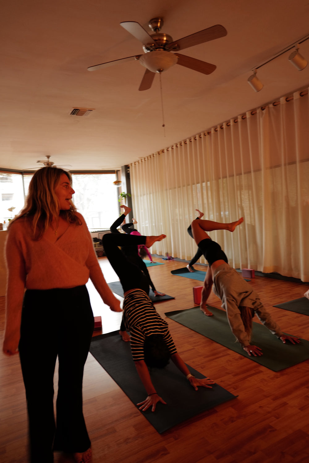 A teacher in an orange top watching a class move through a lunge, ceiling fan overhead and daylight from a side window
